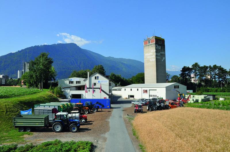 Das Areal an der Rheinmühle in Chur mit Wohnhaus, Silo-Turm und Halle, davor ein Traktor mit Anhänger und ein blauer Container vor Bergen