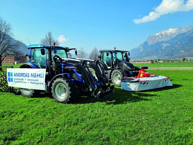 Zwei Valtra-Traktoren mit Frontlader und Mähwerk auf einer Wiese, davor ein Banner von Andreas Mehli
